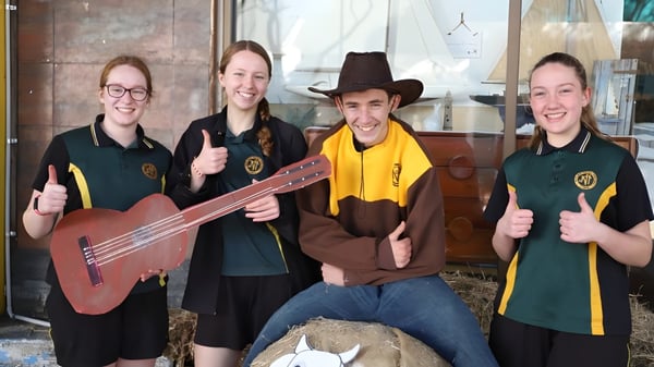 Eine Gruppe von Schülerinnen und Schülern der James Nash State High School steht mit Musikinstrumenten vor einem Holzgebäude.