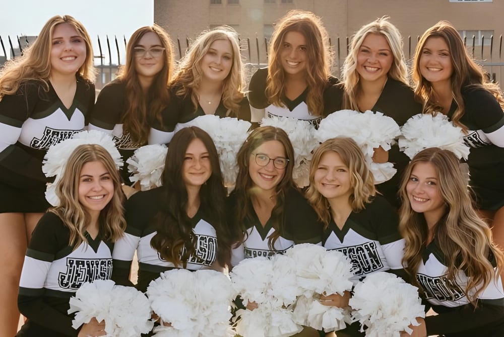 Eine Gruppe junger Schülerinnen in Cheerleader-Uniformen mit Pompons vor einem Gebäude der Jenison High School.
