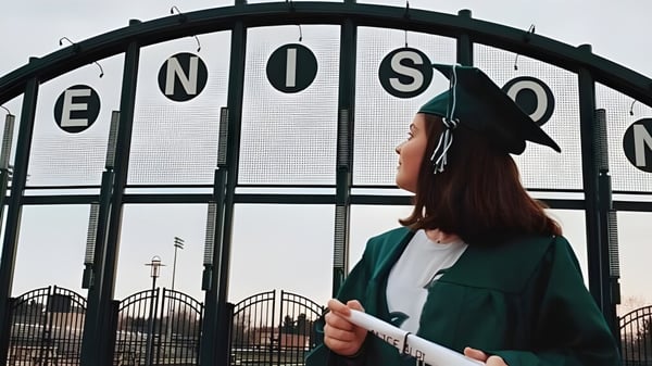 Eine Person mit grünem Mantel steht vor dem großen Metalltor mit dem Schriftzug ENOSC auf dem Gelände der Jenison High School.
