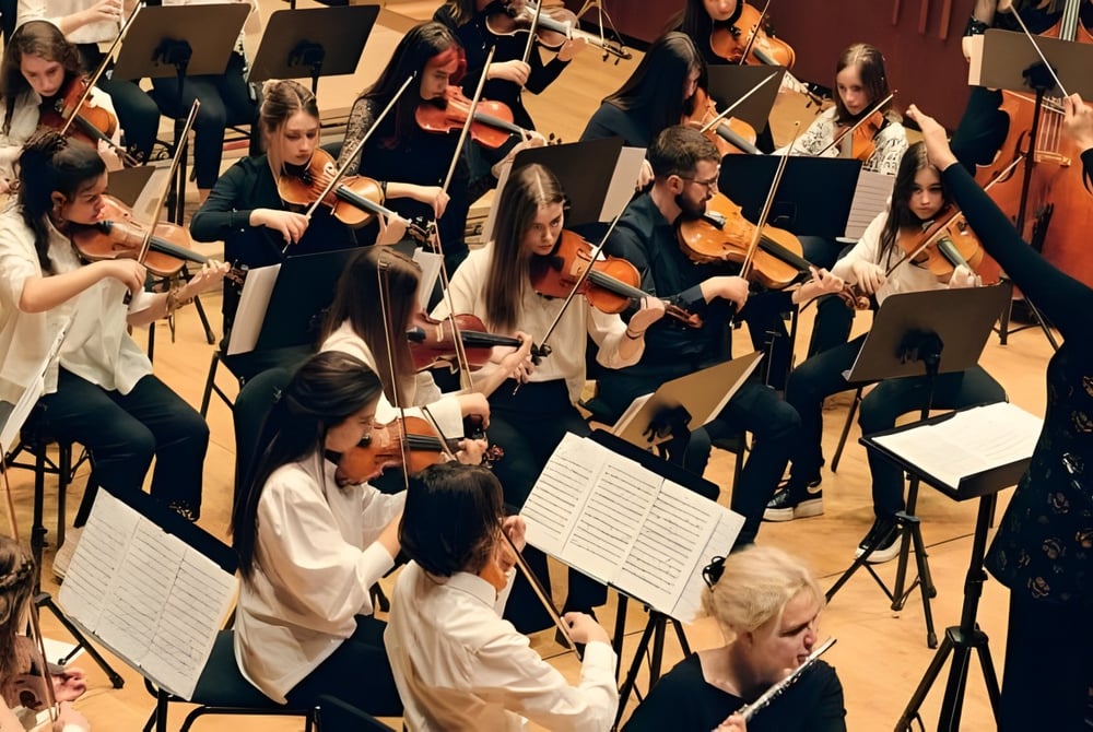 Ein großes Orchester tritt auf der Bühne der Jesus and Mary Secondary School in formeller Kleidung auf und spielt verschiedene Saiteninstrumente.