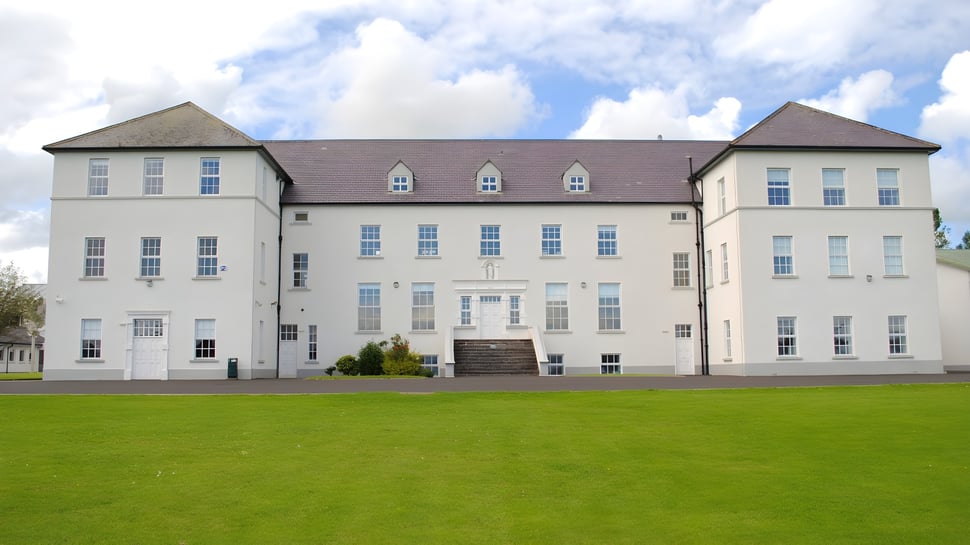 Das weiße Hauptgebäude der Jesus and Mary Secondary School steht auf einem großen Rasenfeld unter bewölktem Himmel.