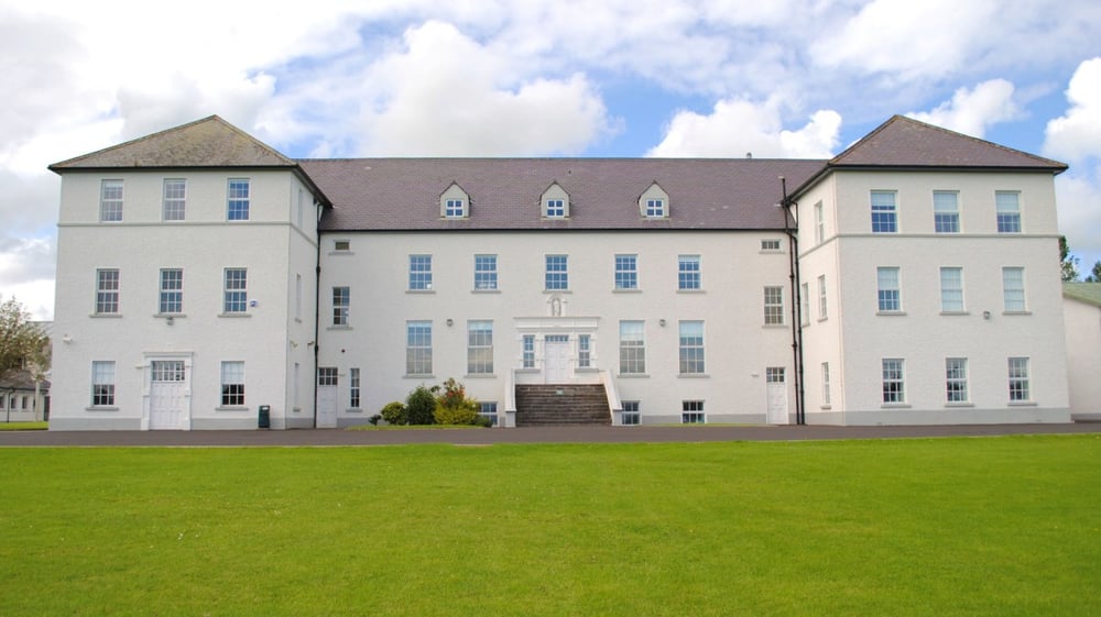 Das große weiße Schulgebäude der Jesus and Mary Secondary School steht auf einer Wiese unter einem bewölkten Himmel.