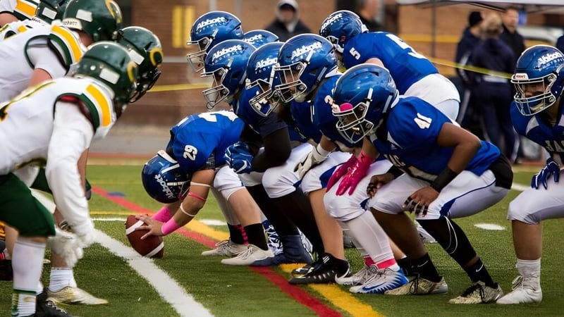 Schüler des John Abbott College stehen in blauen und grünen Trikots auf dem Spielfeld und bereiten sich auf eine Football-Spielzug vor.