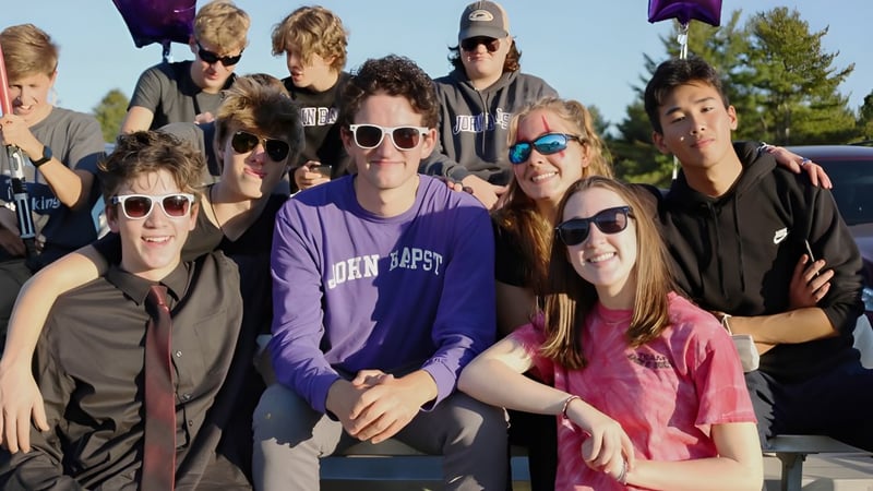 Eine Gruppe von Schülerinnen und Schülern der John Bapst Memorial High School posiert draußen vor buntem Hintergrund aus Bäumen und Himmel.