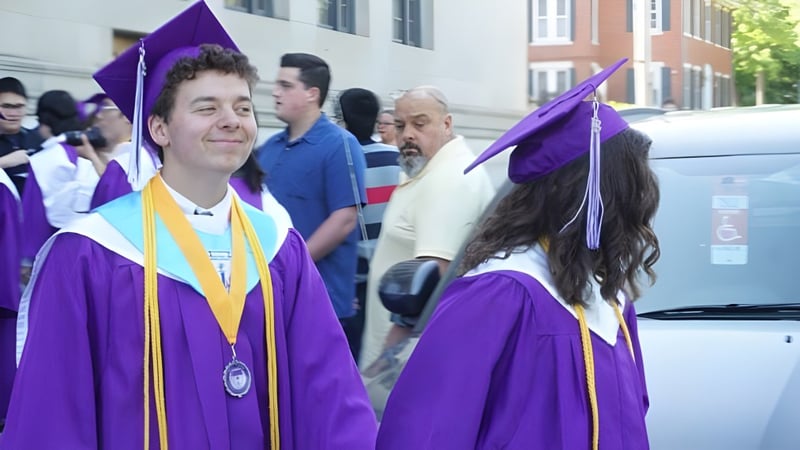 Schüler der John Bapst Memorial High School tragen lila Abschlussroben und Hüte bei einer Abschlussfeier im Freien.