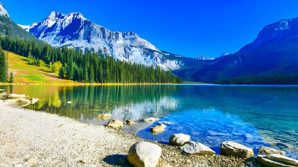 Ein ruhiger alpiner See mit schneebedeckten Bergen und bewaldeter Uferzone in der Nähe der John Barsby Secondary School.