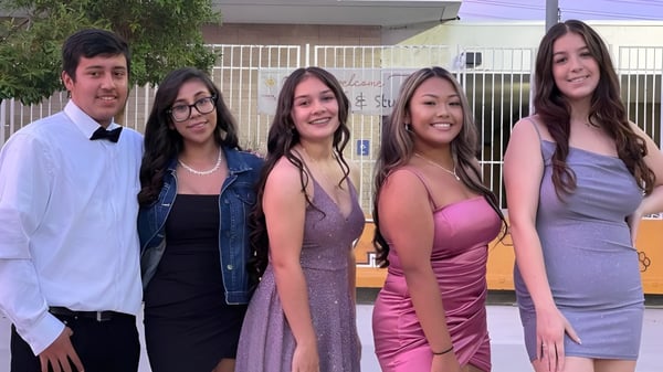 Eine Gruppe Schülerinnen und Schüler steht vor einem Gebäude mit Metallzaun auf dem Gelände der John F. Kennedy High School.