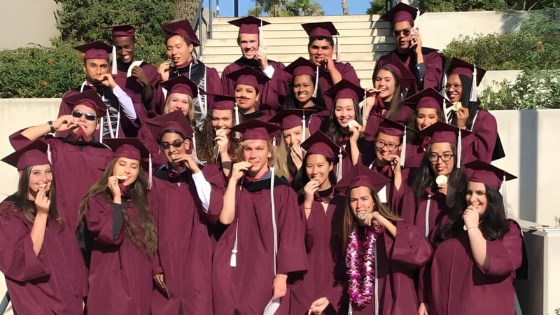 Absolventinnen und Absolventen der John F. Kennedy High School in bordeauxroten Roben stehen vor einem Gebäude.