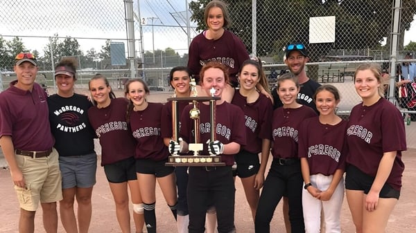 Schülerinnen der John McGregor Secondary School feiern den Gewinn eines Pokals vor einem Sportplatz.