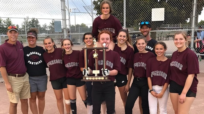 Schülerinnen der John McGregor Secondary School feiern den Gewinn eines Pokals vor einem Sportplatz.