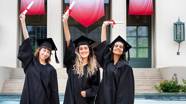 Drei Schülerinnen der John Oliver Secondary School stehen in Abschlusskleidung zusammen und halten stolz ihre Diplome vor dem Schulgebäude mit Pool.