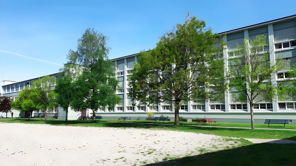 Das mehrstöckige Schulgebäude der John Oliver Secondary School mit grünen Bäumen und einem grasbewachsenen Hof unter blauem Himmel.