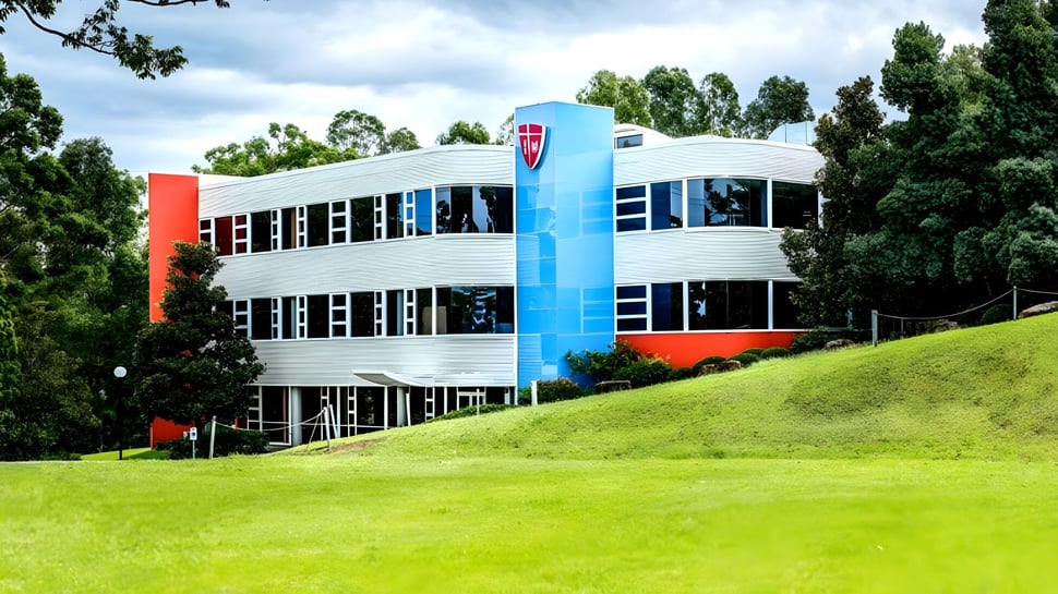 Das moderne Gebäude des John Paul College mit großen Fenstern steht auf einem grasbewachsenen Hügel, umgeben von Bäumen und Grünfläche.