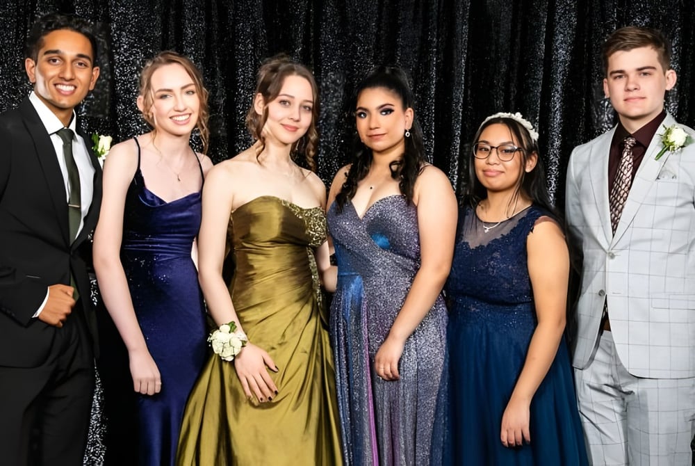 Sechs gut gekleidete Personen posieren gemeinsam vor einem schwarzen Hintergrund auf dem Campus des John Paul College.