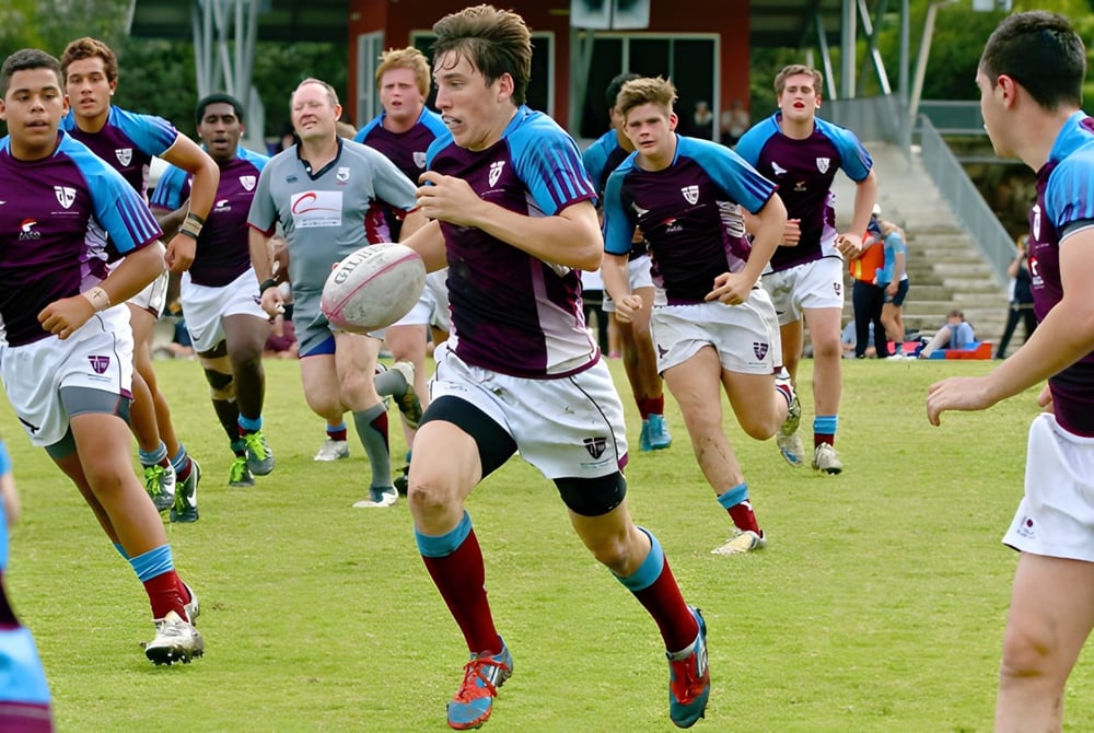 Schüler des John Paul College spielen Fußball auf einem grasbewachsenen Spielfeld vor einem Schulgebäude.