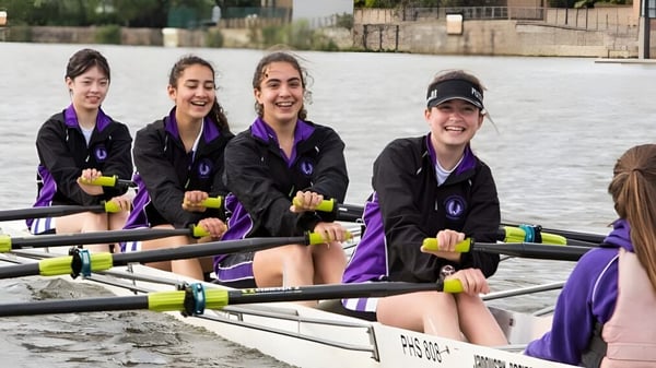 Eine Gruppe Schülerinnen der John Scottus Secondary School sitzt in einem Ruderboot auf dem Wasser mit Stadtblick im Hintergrund.