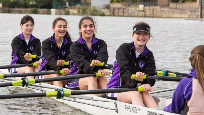 Eine Gruppe Schülerinnen der John Scottus Secondary School sitzt in einem Ruderboot auf dem Wasser vor einer Stadtansicht.