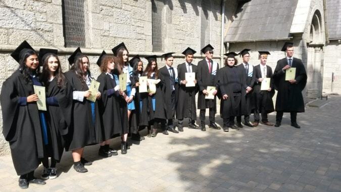 Eine Gruppe von Absolventinnen und Absolventen der John Scottus Secondary School in akademischer Kleidung steht vor einem steinernen Gebäude mit Rundbogenfenstern.