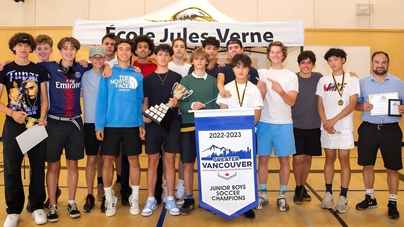 Eine Gruppe von Schülerinnen und Schülern der Jules Verne school Vancouver posiert in der Sporthalle mit einem Schulbanner und einer Meisterschaftstafel.