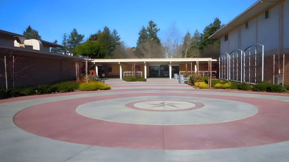 Der gepflasterte Innenhof mit rundem Muster auf dem Campus der Justin Siena High School ist von Gebäuden mit großen Fenstern und Kiefern umgeben.