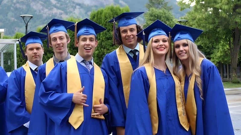 Absolventen der J.V. Humphries Secondary School stehen in ihren blauen und goldenen Talaren gemeinsam im Freien vor Bäumen und Bergen.