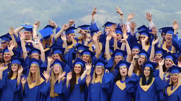 Absolventen der Kalamalka Secondary School feiern in blau-gelben Talaren ihre Graduierung vor bergiger Landschaft.