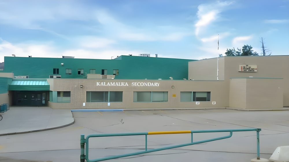 Das mehrstöckige grün-beige Gebäude der Kalamalka Secondary School mit sichtbarem Schriftzug und gepflastertem Bereich im Vordergrund.