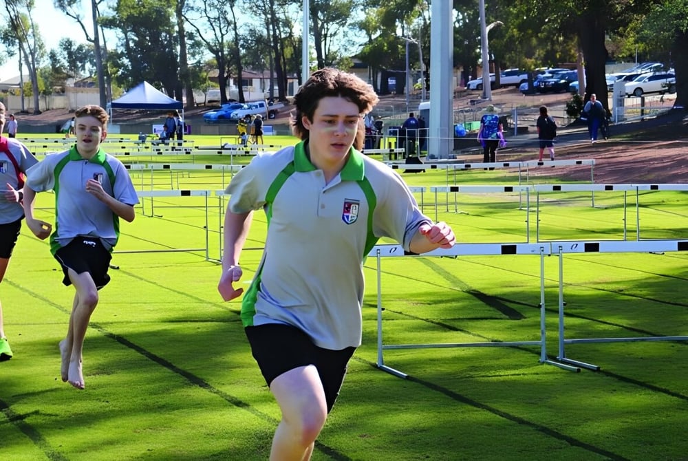 Eine Person in weißer Sportkleidung läuft auf dem Sportplatz der Kalamunda Senior High School über Hürden auf einem Rasenfeld.