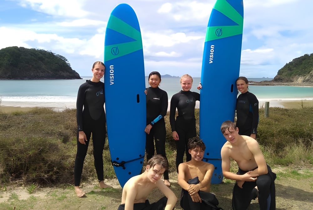 Schüler der Kamo High School stehen in Neoprenanzügen neben großen Surfboards am Strand mit Bergen und Wasser im Hintergrund.