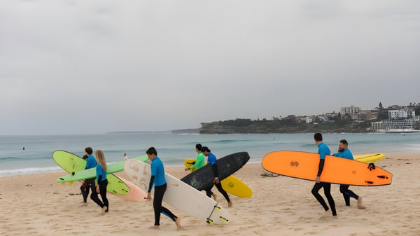 Eine Gruppe von Schülerinnen und Schülern der Keebra Park State High School trägt bunte Surfboards am Strand mit der Stadt im Hintergrund.