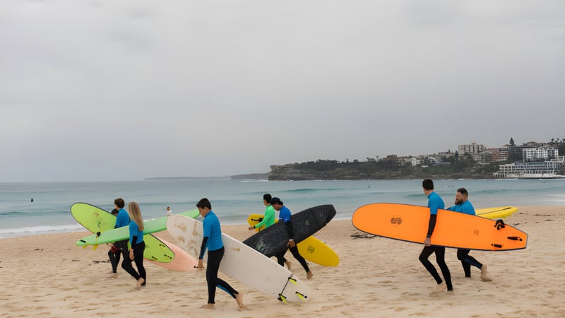 Eine Gruppe von Schülerinnen und Schülern der Keebra Park State High School trägt bunte Surfboards am Strand mit der Stadt im Hintergrund.