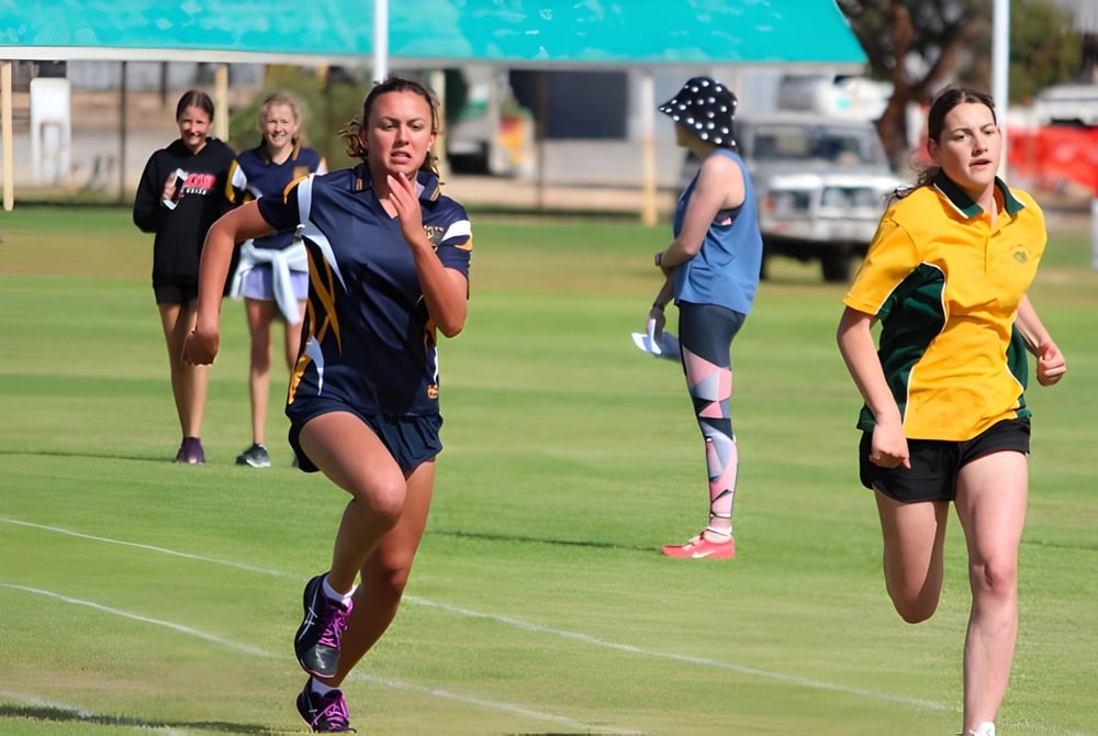 Mehrere Athletinnen laufen auf einem Grasfeld vor Gebäuden auf dem Gelände der Keith Area School.