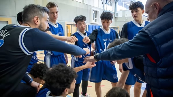 Eine Gruppe junger Basketballspieler in blauen Trikots hört auf die Anweisungen des Trainers auf dem Gelände der Kells Academy.