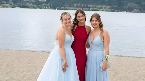 Drei Schülerinnen der Kelowna Secondary School stehen in formellen Kleidern am Strand mit See und Bergen im Hintergrund.