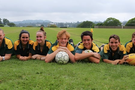 Eine Gruppe junger Fußballspielerinnen sitzt auf einem grasbewachsenen Spielfeld der Kenmore State High School, umgeben von Bäumen und wolkigem Himmel.