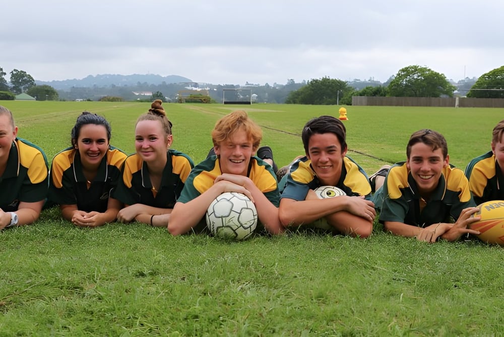 Eine Gruppe junger Fußballspielerinnen sitzt auf einem grasbewachsenen Spielfeld der Kenmore State High School, umgeben von Bäumen und wolkigem Himmel.