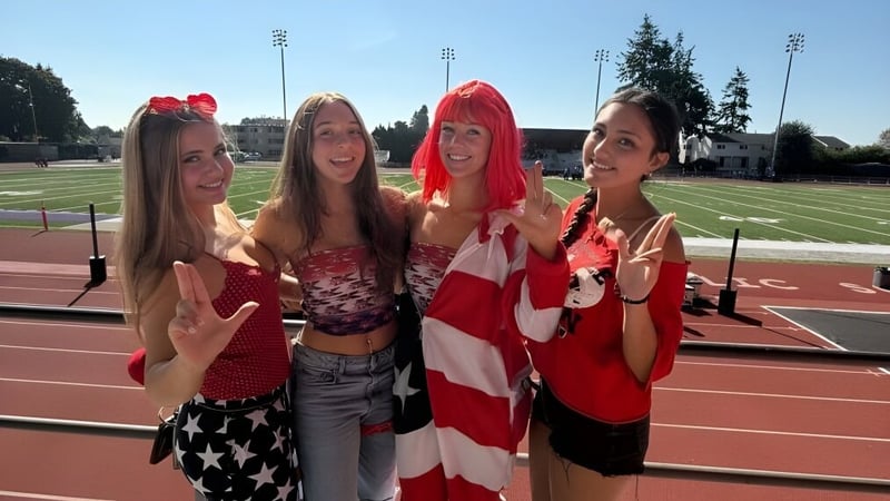 Vier Schülerinnen der Kennedy Catholic High School posieren in patriotischer Kleidung auf einem Sportfeld mit Stadion im Hintergrund.