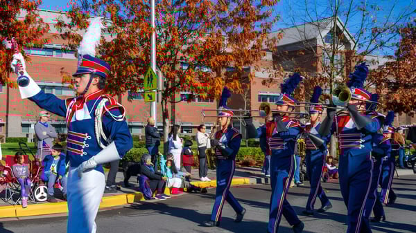 Mitglieder der Marching Band der Kennedy Catholic High School treten in bunten Uniformen auf einer von Herbstbäumen gesäumten Straße auf.