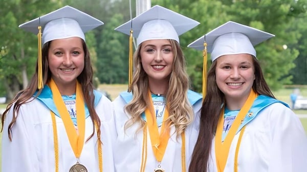 Drei Schülerinnen der Kenston Forest School stehen lächelnd in ihren Abschlussroben vor grünem Hintergrund.