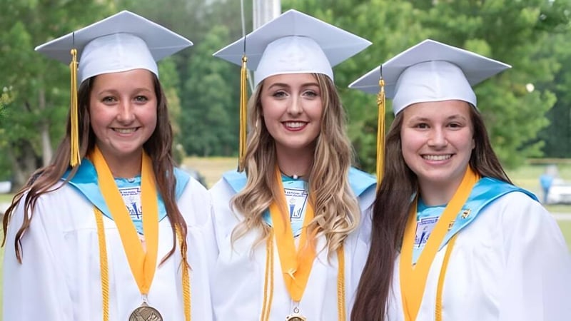 Drei Schülerinnen der Kenston Forest School stehen lächelnd in ihren Abschlussroben vor grünem Hintergrund.