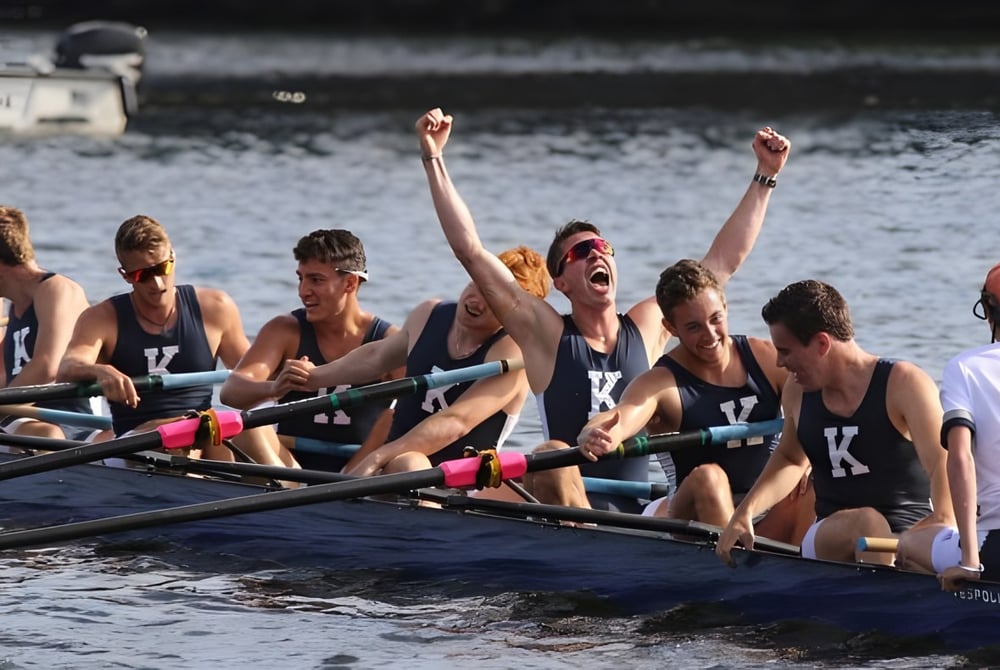 Die Rudermannschaft der Kent School feiert gemeinsam in ihrem Boot auf dem Wasser vor einer Stadtkulisse.