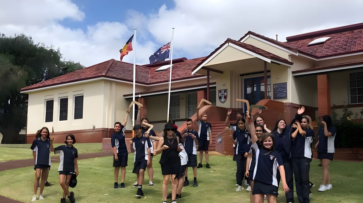 Eine Gruppe Schülerinnen und Schüler in Schuluniformen steht vor dem traditionellen Gebäude der Kent Street Senior High School mit wehenden Flaggen.