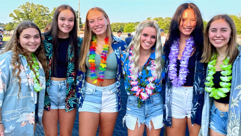 Eine Gruppe junger Schülerinnen der Kettle Moraine High School in farbenfroher Festkleidung posiert draußen vor Bäumen und klarem Himmel.