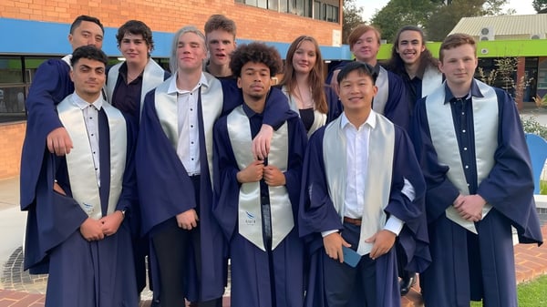 Eine Gruppe von Schülerinnen und Schülern des Kiara College steht in ihren Abschlussroben vor einem Backsteingebäude.