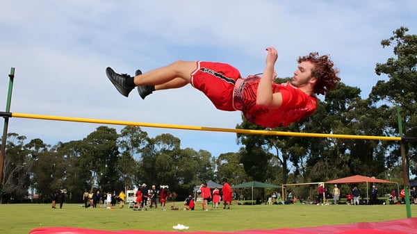 Eine Schülerin des Kiara College springt im Freien über eine Hochsprunglatte vor Bäumen und weiteren Personen.