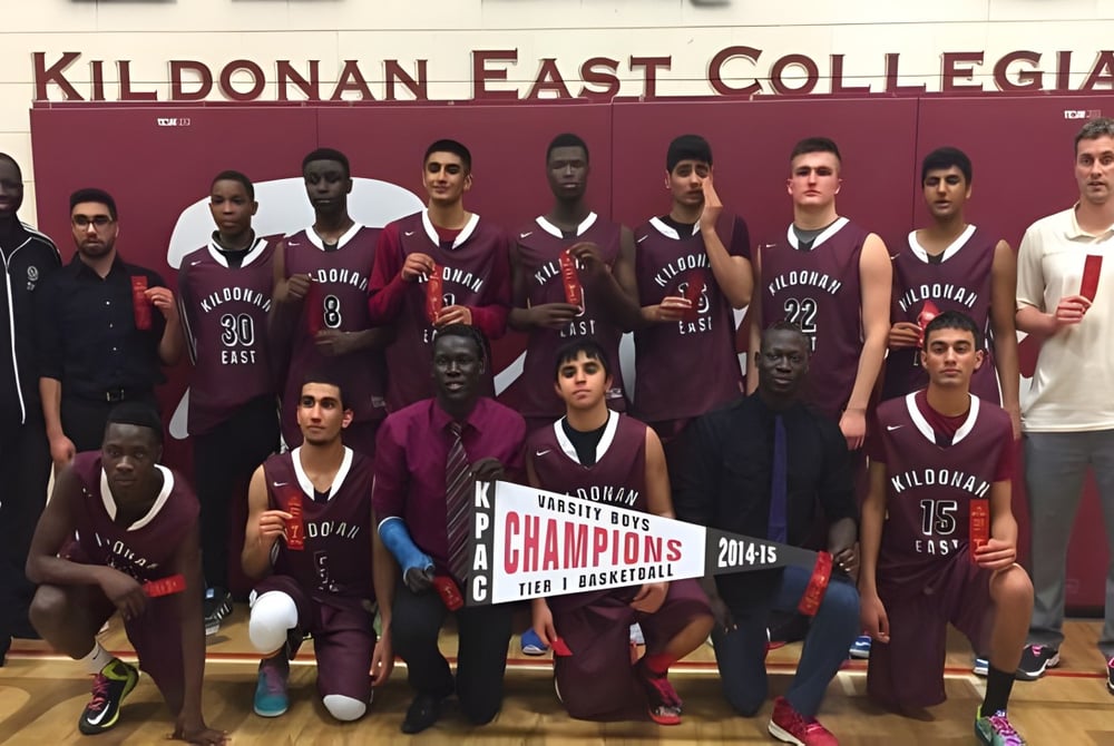 Das Basketballteam des Kildonan East Collegiate posiert gemeinsam mit einem Pokal vor einem Schild der Schule.