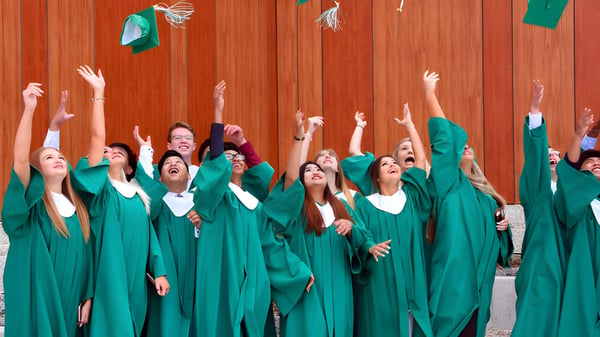 Absolventinnen und Absolventen der Kildonan East Collegiate in grünen Talaren feiern vor einer Holzwand.