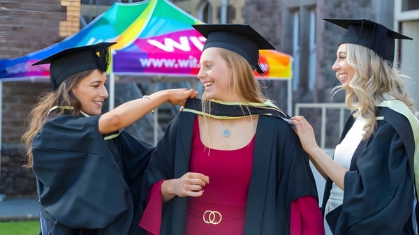 Drei Schülerinnen der Kilkenny City Vocational School feiern ihren Abschluss vor einem bunten Hintergrund mit einem WIN-Schild.