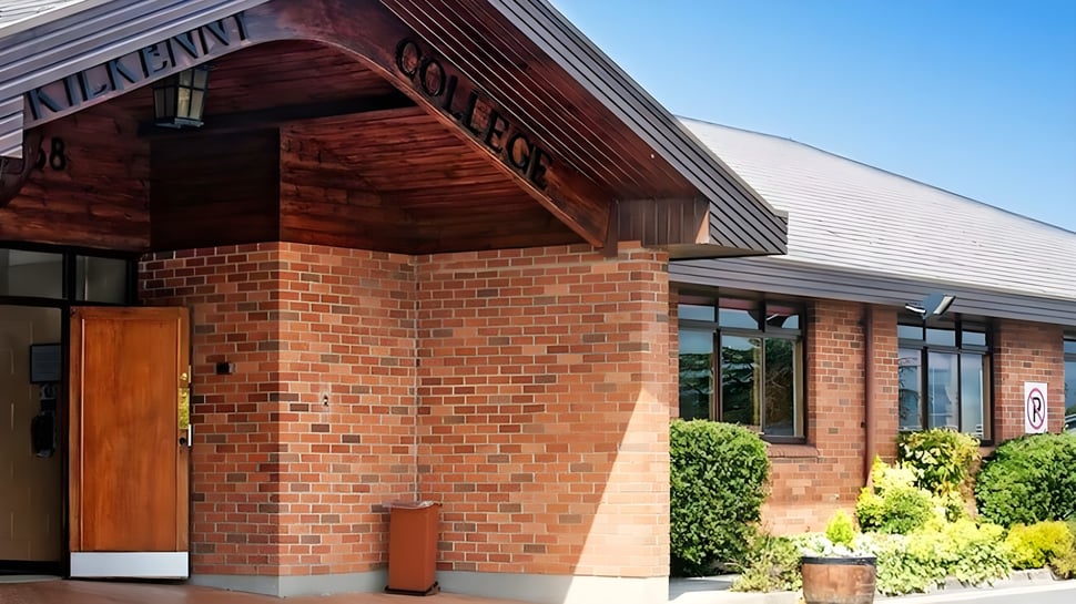 Das Backsteingebäude mit einem hölzernen Vordach des Kilkenny College ist von Grün umgeben unter blauem Himmel.