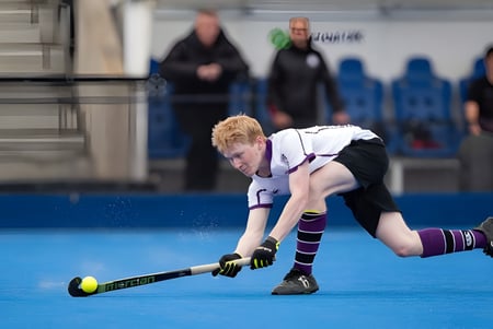 Eine Person spielt Feldhockey auf dem blauen Kunstrasenplatz der Kimbolton School vor einer Gruppe von Zuschauern.